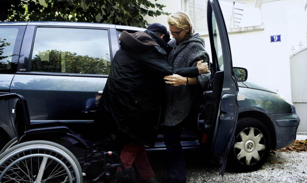Personne en fauteuil roulant assistée par un aidant familial pour monter dans une voiture, soulignant le soutien quotidien et la mobilité des personnes dépendantes