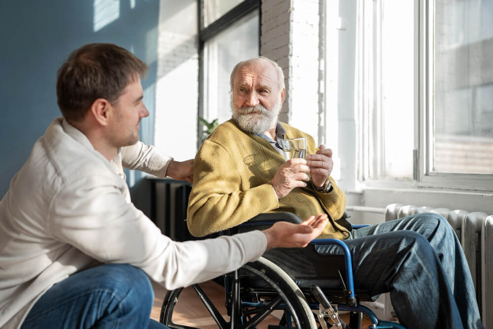 Jeune homme attentionné parlant à un homme âgé barbu en fauteuil roulant, qui tient un verre d'eau, illustrant le rôle de l'aidant familial dans le quotidien des seniors dépendants