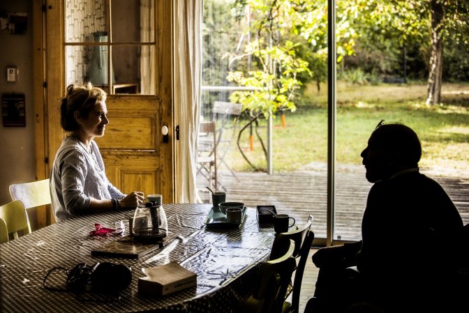 Deux personnes assises à une table ensoleillée près d'une grande fenêtre, une femme souriante en pleine lumière et un homme en silhouette, évoquant la discussion et l'échange au sein d'une relation d'aide