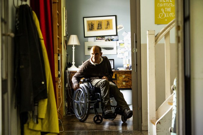 Homme en fauteuil roulant se déplaçant dans un couloir lumineux de sa maison, illustrant l'autonomie et les défis de la mobilité pour les personnes dépendantes